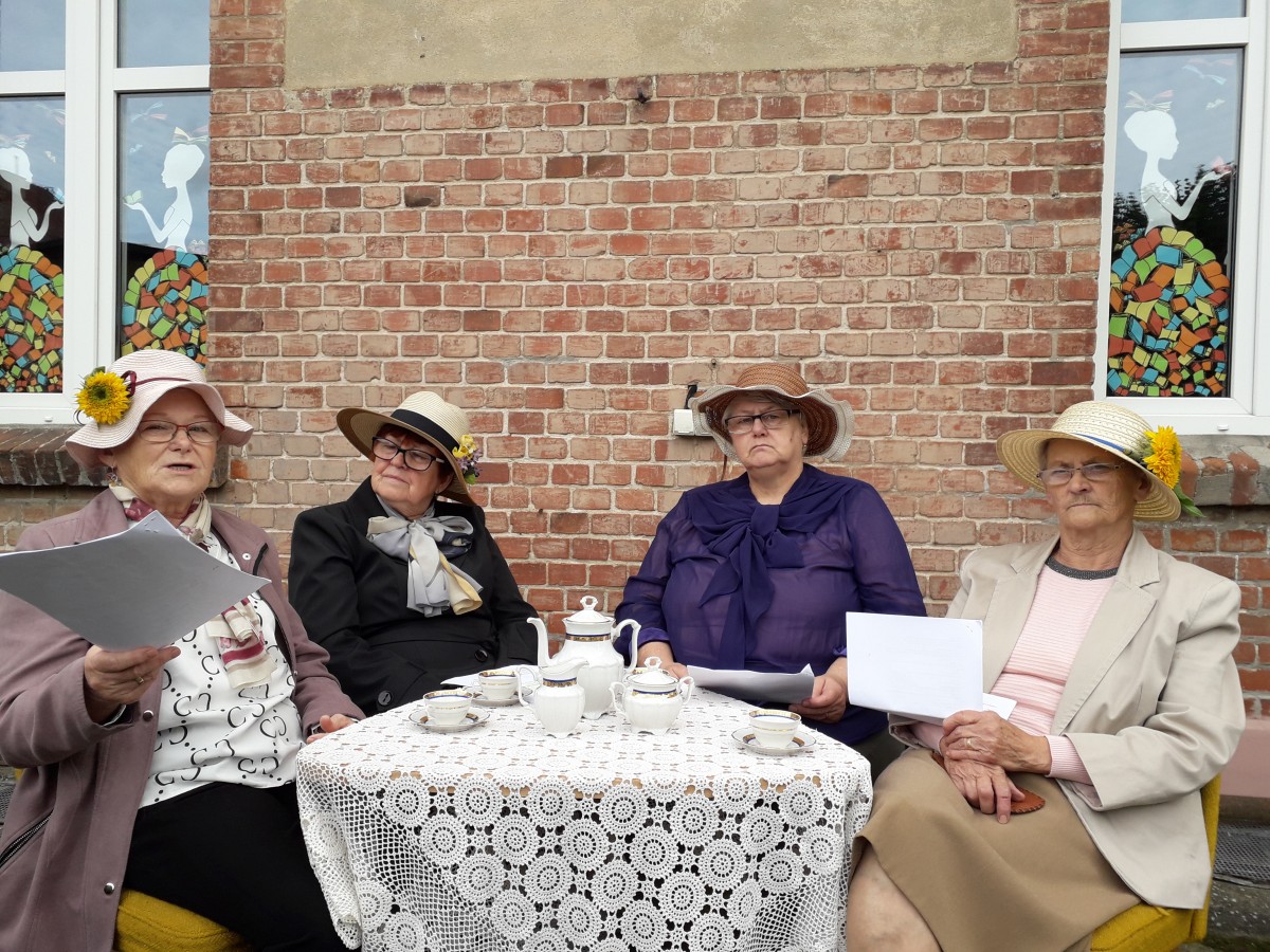 cztery kobiety siedzą w kapeluszach przy stoliku, serwis kawowy, w tle ściana z cegieł