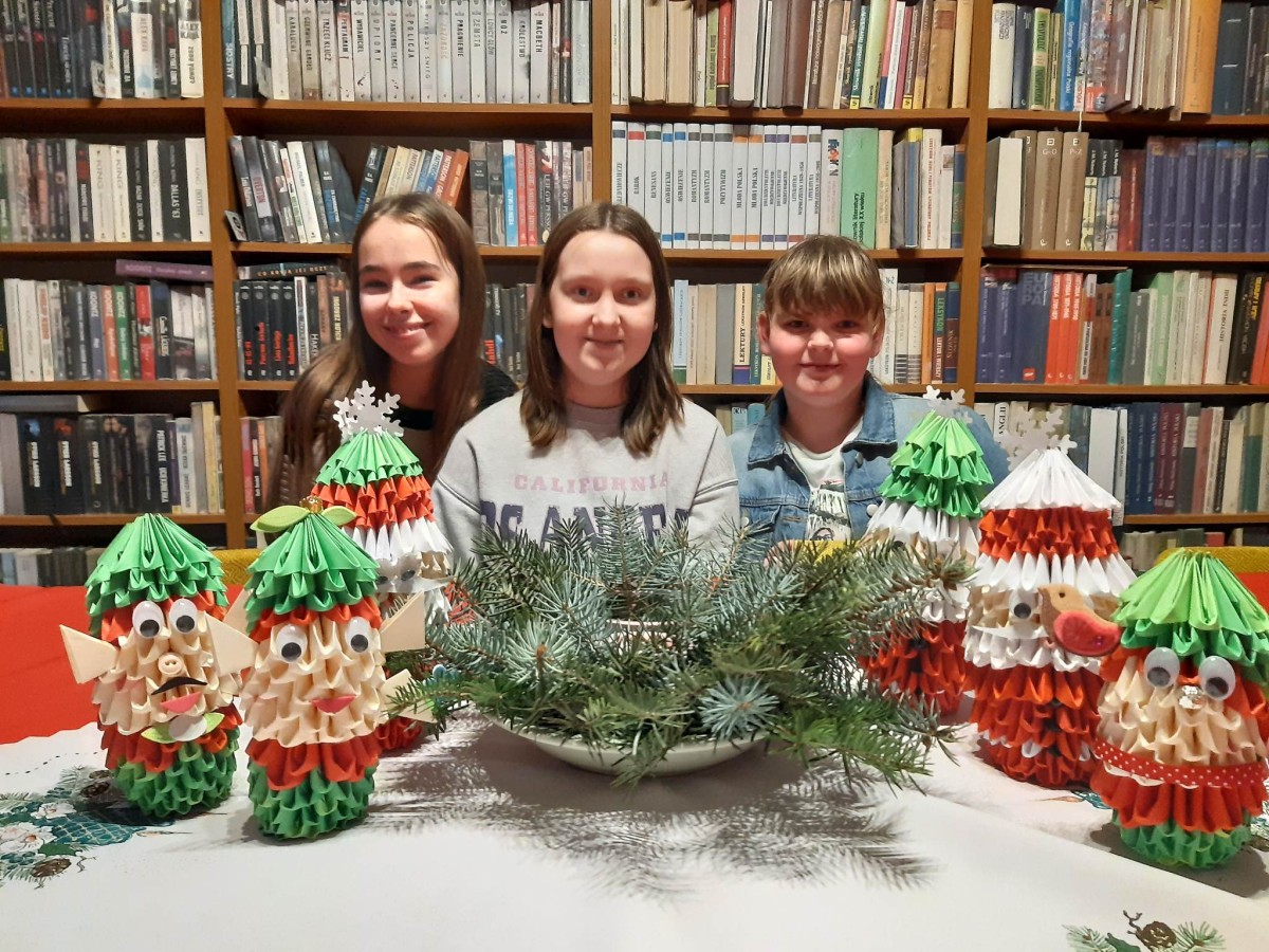 zdjęcie na tle regałów z książkami, trzy dziewczynki, stół, stroik świąteczny, mikołaje i elfy z origami